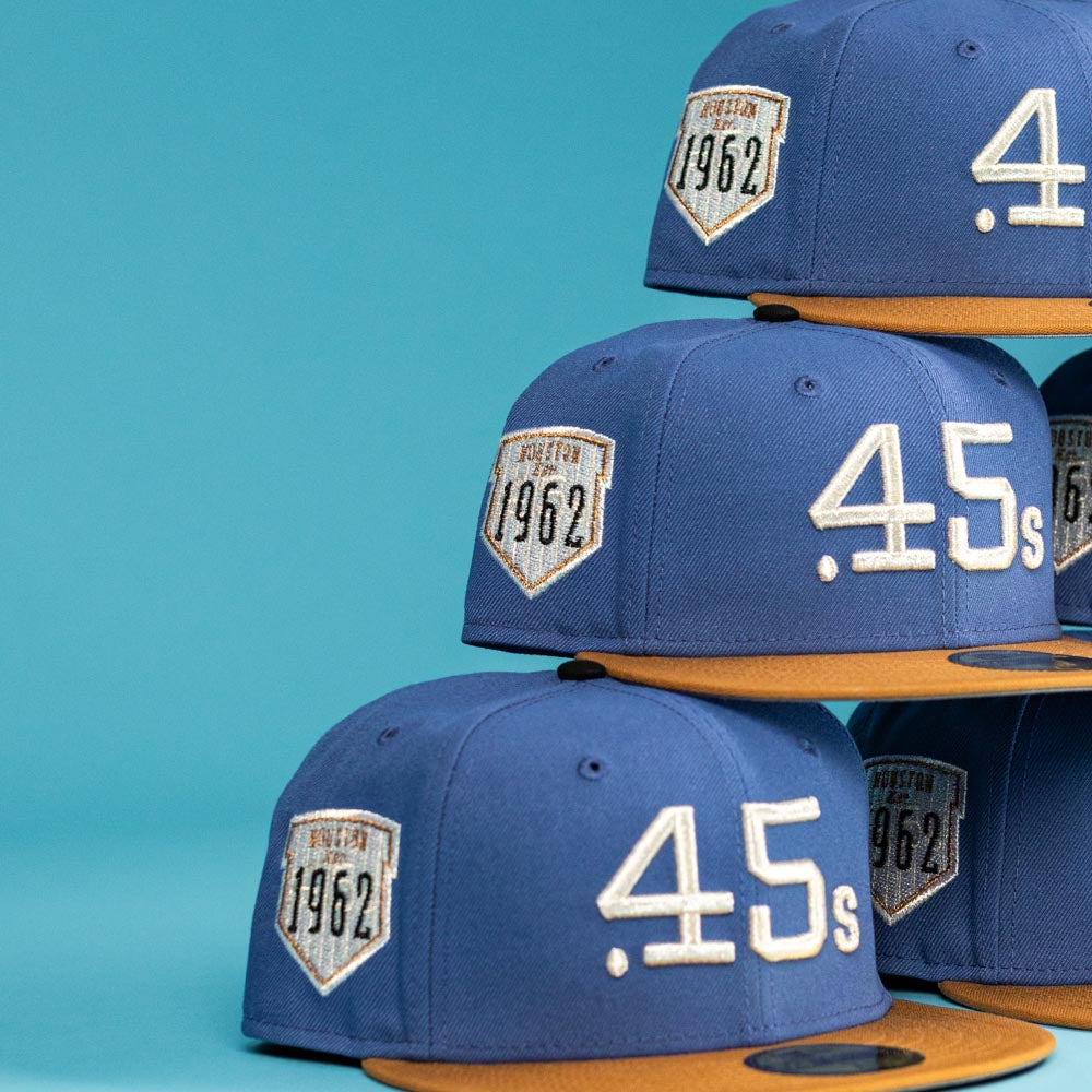 A stack of Harbor Mark Canvas Houston Astros New Era Hat 5950 Fitted caps with gold brims, featuring ".45s" on the front and a Houston 1962 patch on the side, displayed against a blue background.