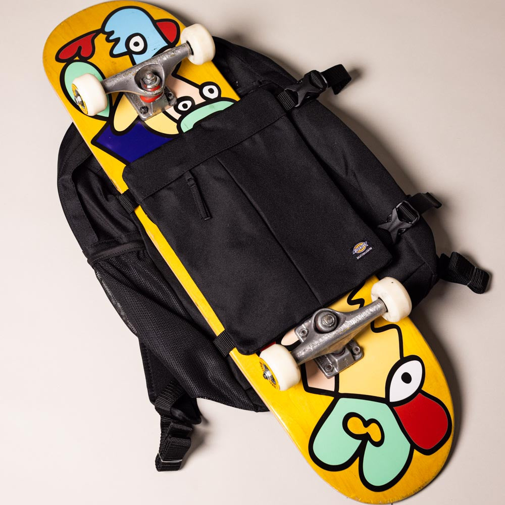 The A8675BLK Black Mike Anderson Pro Skate Dickies Skateboard Strap Backpack holds a yellow skateboard with a vibrant cartoon face graphic in blue, red, green, and beige, photographed from above on a light background.