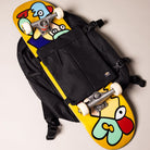 The A8675BLK Black Mike Anderson Pro Skate Dickies Skateboard Strap Backpack holds a yellow skateboard with a vibrant cartoon face graphic in blue, red, green, and beige, photographed from above on a light background.