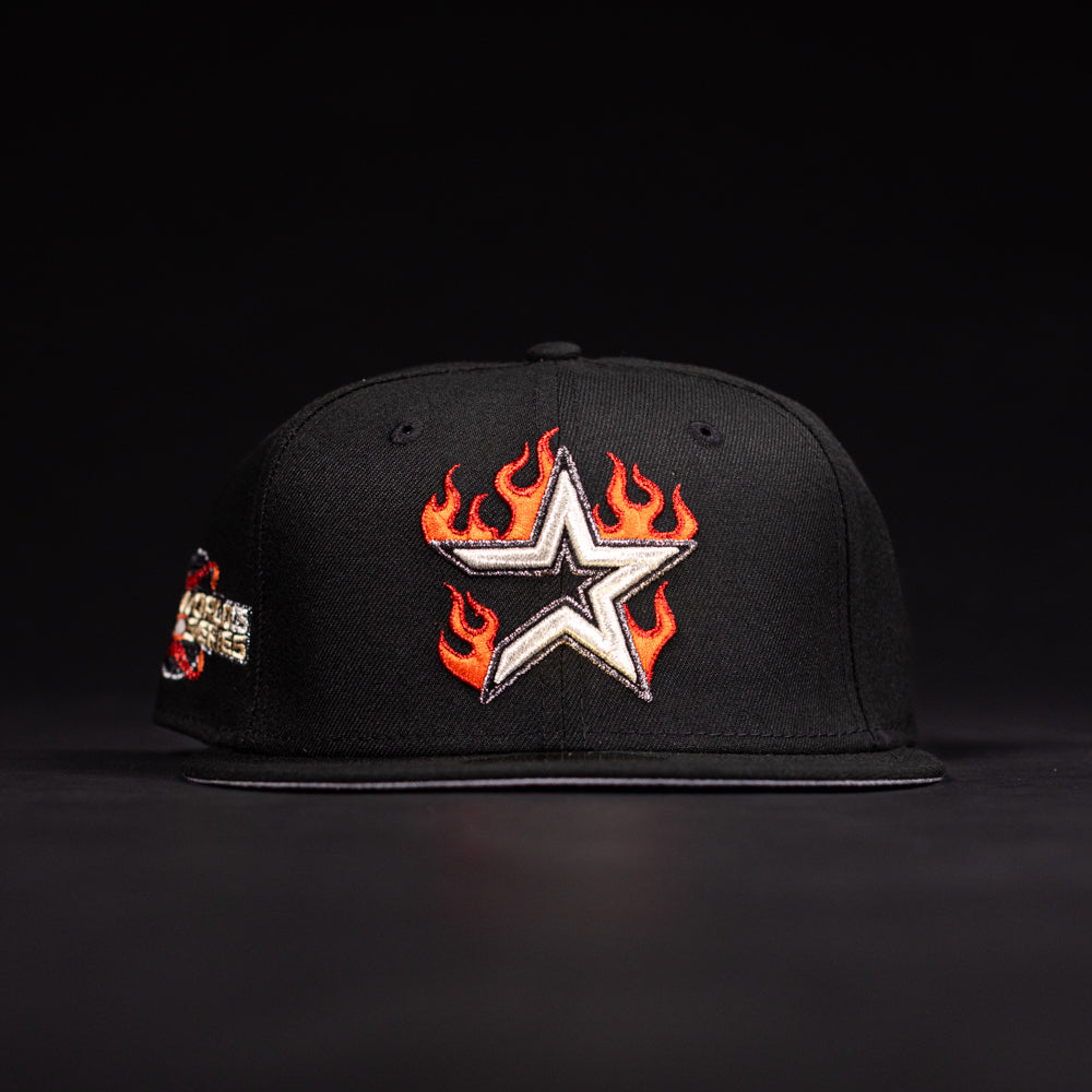 The La Flame Rushmore WS05 Fire Black Houston Astros New Era 5950 Fitted Hat features a white star with orange flames on the front, a colorful side patch, and is shown against a dark background.