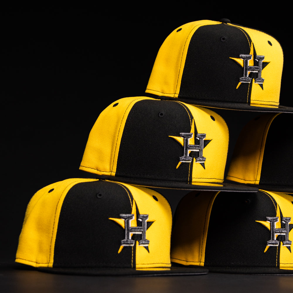 Four Hazmat Rushmore Gold Black Pinwheel Houston Astros New Era Hat 5950 Fitted caps, featuring a stylized H logo, are stacked atop each other against a black background.