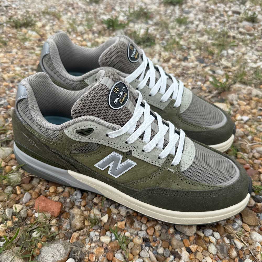 A pair of 933 JKL Andrew Reynolds Grey Day New Balance Numeric Skateboarding Shoes Suede with white laces rest on gravel with grass nearby, highlighting their stylish, durable design for skateboarding.