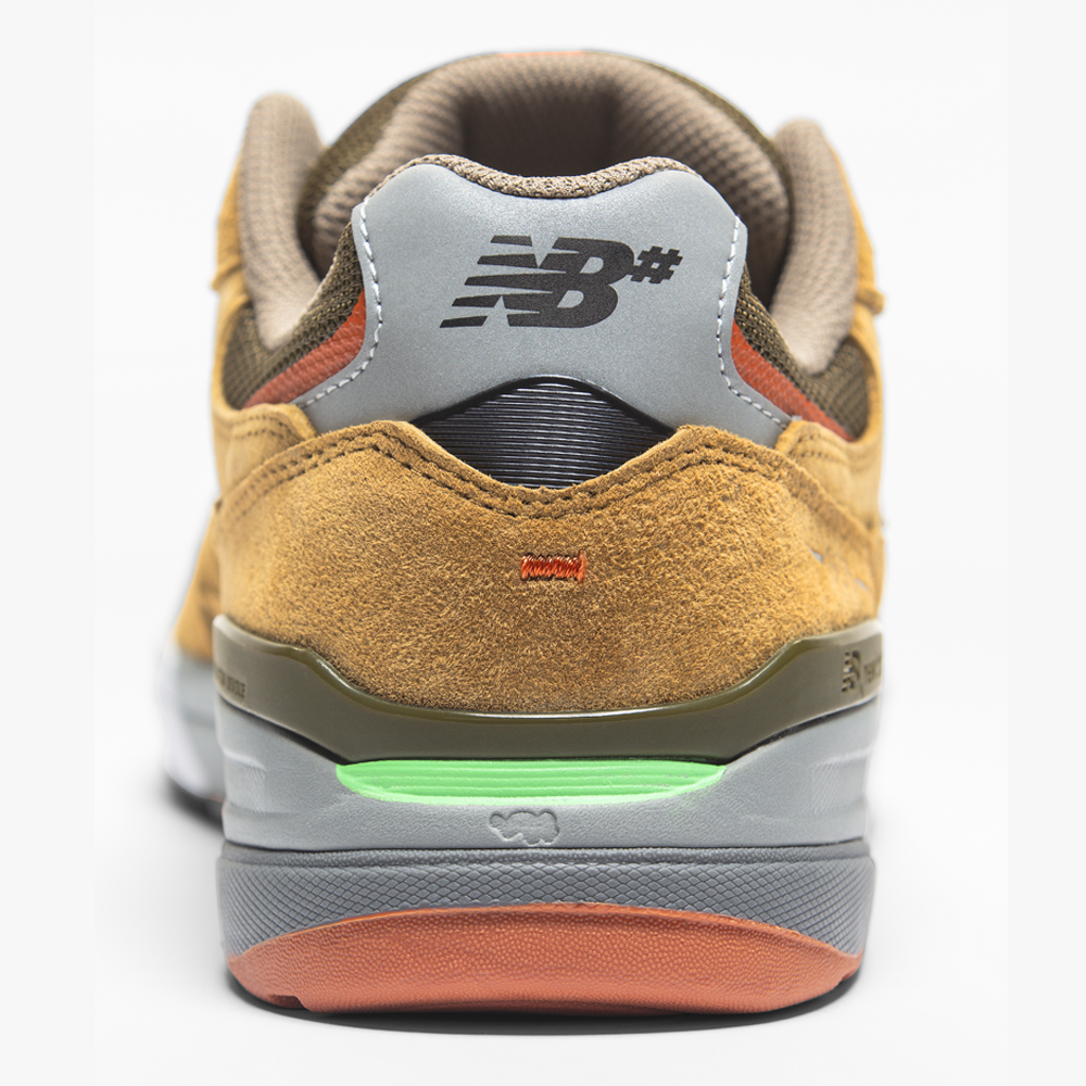 A close-up of the 933 MNO Andrew Reynolds Brown Grey New Balance Numeric suede skateboarding shoe, featuring a gray heel tab, black NB logo, and green and orange accents on the midsole and outsole.