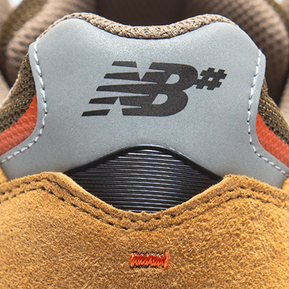 Close-up of the heel of the 933 MNO Andrew Reynolds Brown Grey New Balance Numeric skate shoe, showing a grey panel with a black NB# logo, tan suede material, and orange stitching.