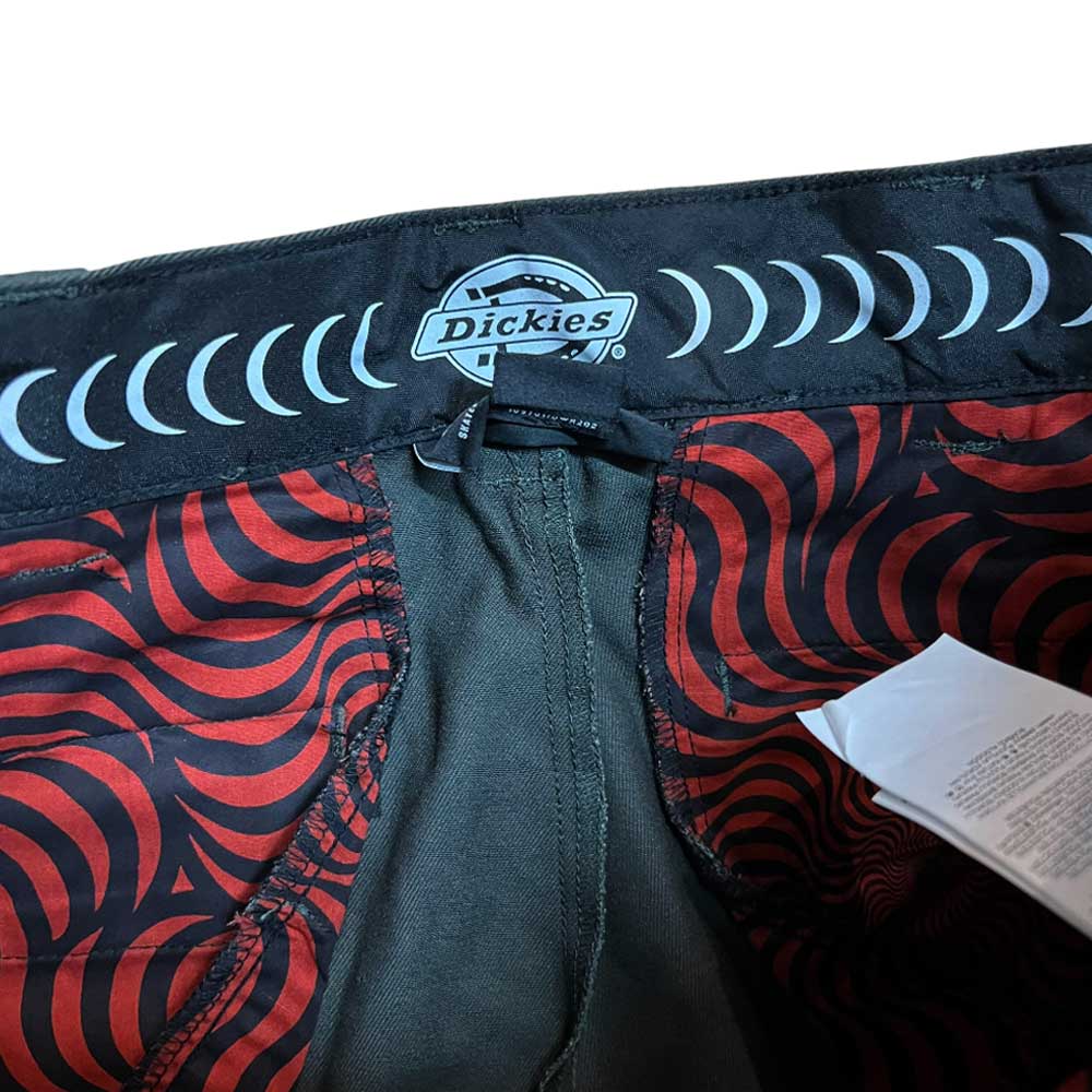 The A865R0OG Olive Spitfire Double Knee Dickies Skateboarding Pant features a waistband with the Dickies logo and crescent pattern, plus a red and black geometric inner lining. A white care tag is partly visible near the Double Knee area.