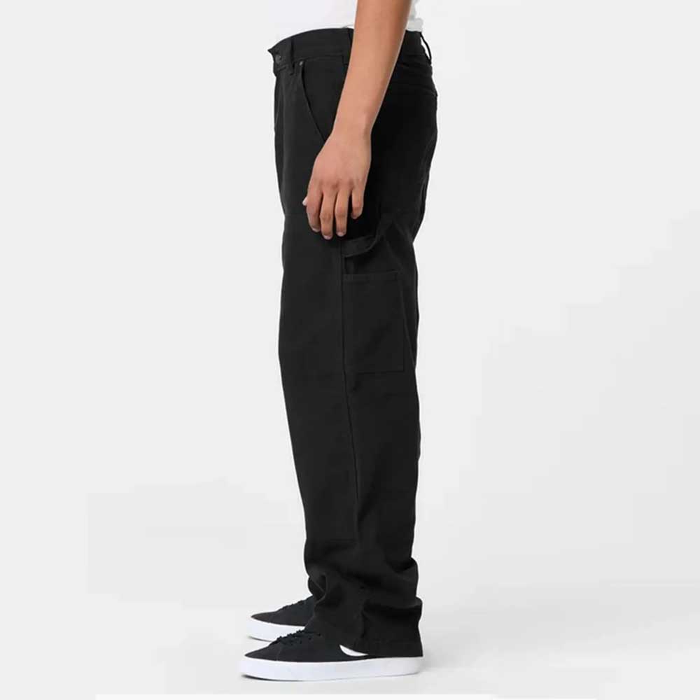 A person stands sideways against a plain light background, showing only the lower body, wearing A87PE Black Double Knee Canvas Carpenter Pant Dickies Skateboarding and black sneakers with white soles.