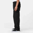 A person stands sideways against a plain light background, showing only the lower body, wearing A87PE Black Double Knee Canvas Carpenter Pant Dickies Skateboarding and black sneakers with white soles.