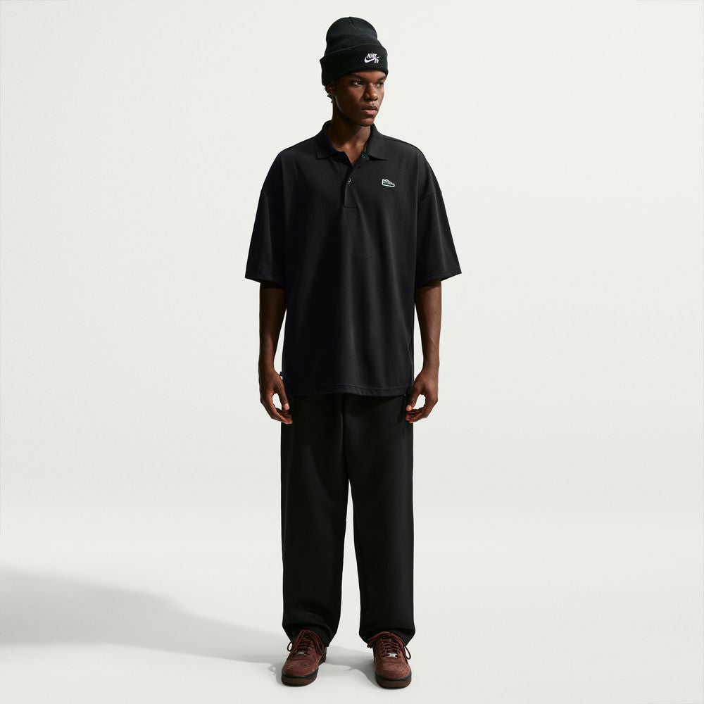 A person wears a loose-fitting black AF1 Black Court Green Solid Break Polo IV0597010 Polo Top Nike SB Skateboarding, black trousers, a black beanie, and dark sneakers.