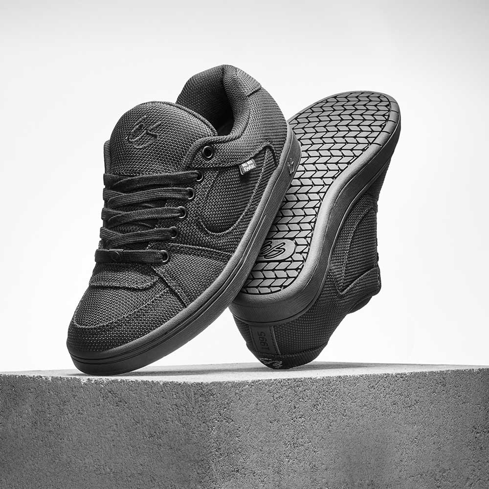 A pair of Accel Kevlar Black Es Skateboarding Shoes are shown on a concrete block, one upright and one tilted to reveal the textured sole—ideal for skateboarding.