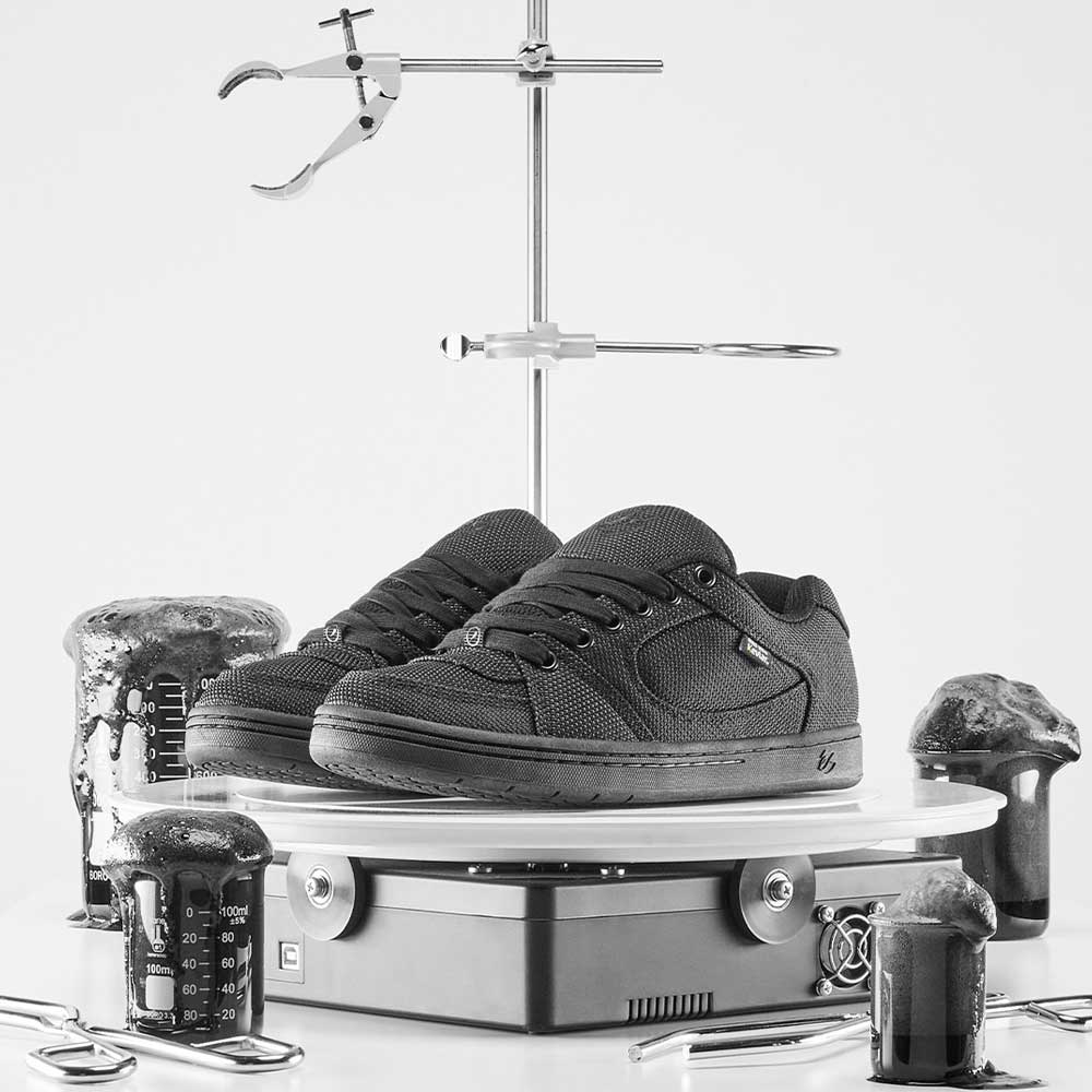 A pair of Accel Kevlar Black Es Shoes for skateboarding sits on a lab platform, surrounded by beakers and test tubes with dark foaming liquid, blending skate culture with a bold, science-inspired monochrome aesthetic.