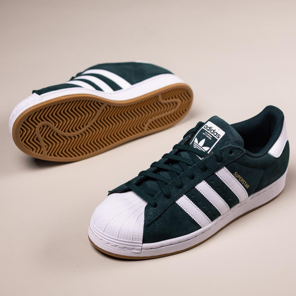 The KI5732 Green White Superstar ADV Adidas Skateboarding Suede Shoes are shown on a light beige surface, with one shoe upright and the other displaying its gum sole. The shoes feature dark green suede, white stripes, and white shell toes.