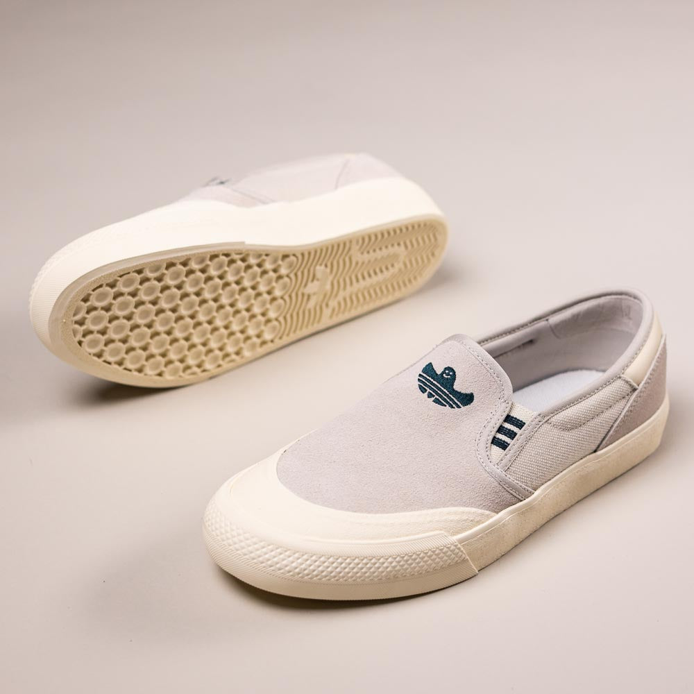 The HQ4721 Gray White Shmoofoil Slip RX Adidas Skateboarding Suede Shoes feature a light grey slip-on design with white rubber soles; one shoe highlights the blue embroidered logo, while the other shows the vulcanised outsole’s textured pattern.