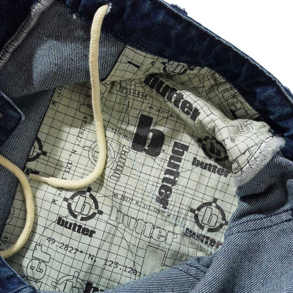 Close-up of Aleka Dark Indigo Denim Jeans Butter Goods, featuring a printed fabric lining with "butter" and repeating b logo over a grid, plus white drawstring and carpenter pocket stitching.