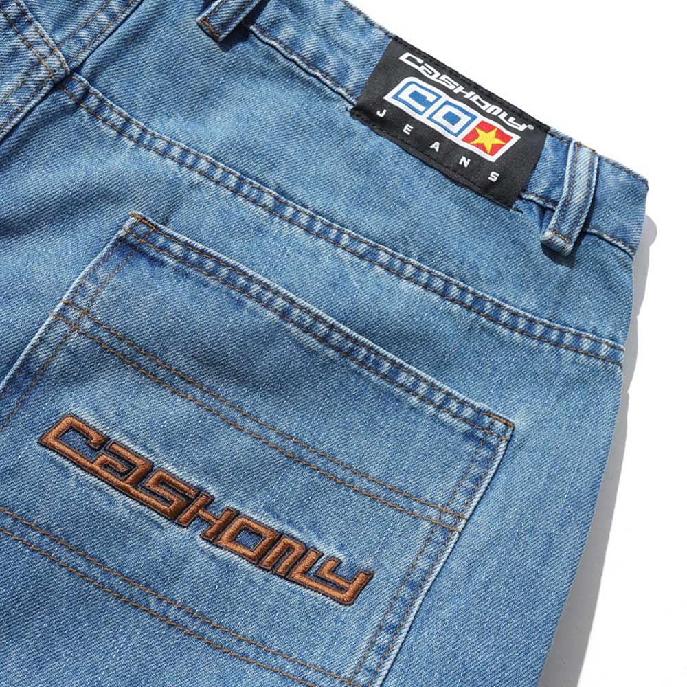 Close-up of Aleka Denim Jeans Distressed Mid Blue (COD153201) showing a back pocket with "CASHONLY" embroidered in brown and a black label reading "CASHONLY CEO JEANS" with a yellow star on a red background above the pocket.