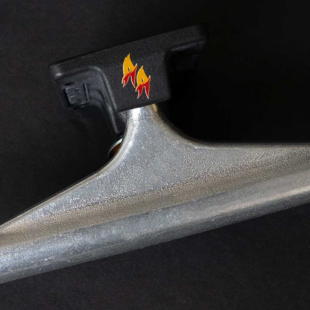 Close-up of an Andrew Allen Hollow Silver Black Standard INDY 159 Stage 11 truck set (two pieces) with a black baseplate and a small yellow and red flame graphic, shown against a dark background.