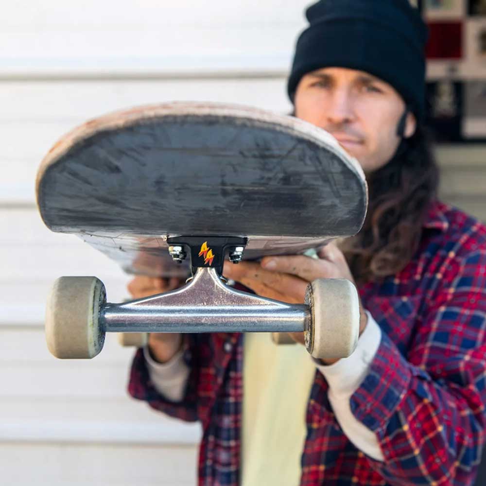 A person in a plaid shirt and black beanie holds a skateboard horizontally, featuring the Andrew Allen Hollow Silver Black Standard INDY 159 Stage 11 Trucks Set of TWO, while their face stays out of focus in the background.