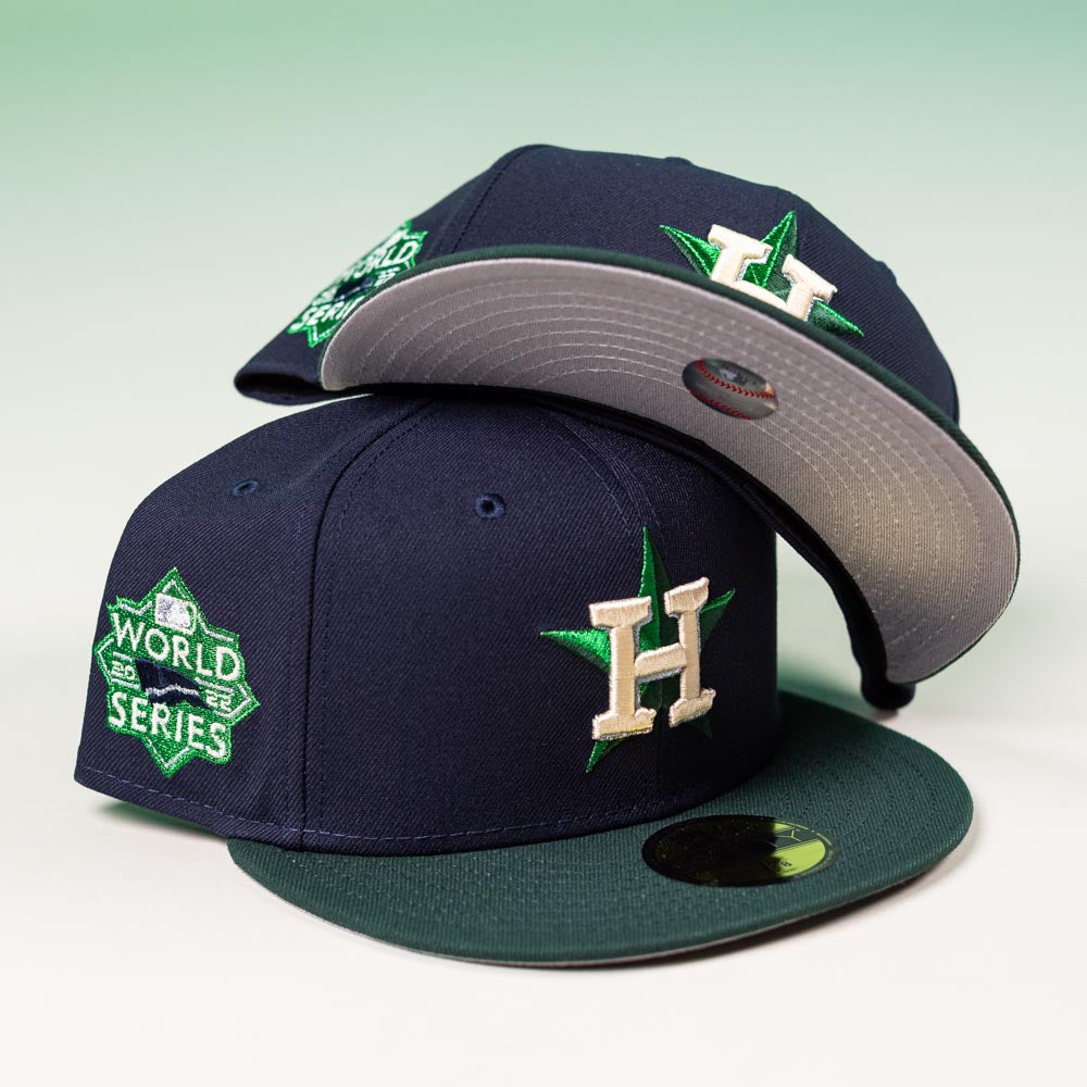 Two Creator Pack Navy Green Houston Astros New Era 5950 Fitted Hats, each with an "H" logo and green star. One features a World Series patch. Both caps are shown on a light surface with a gradient background.
