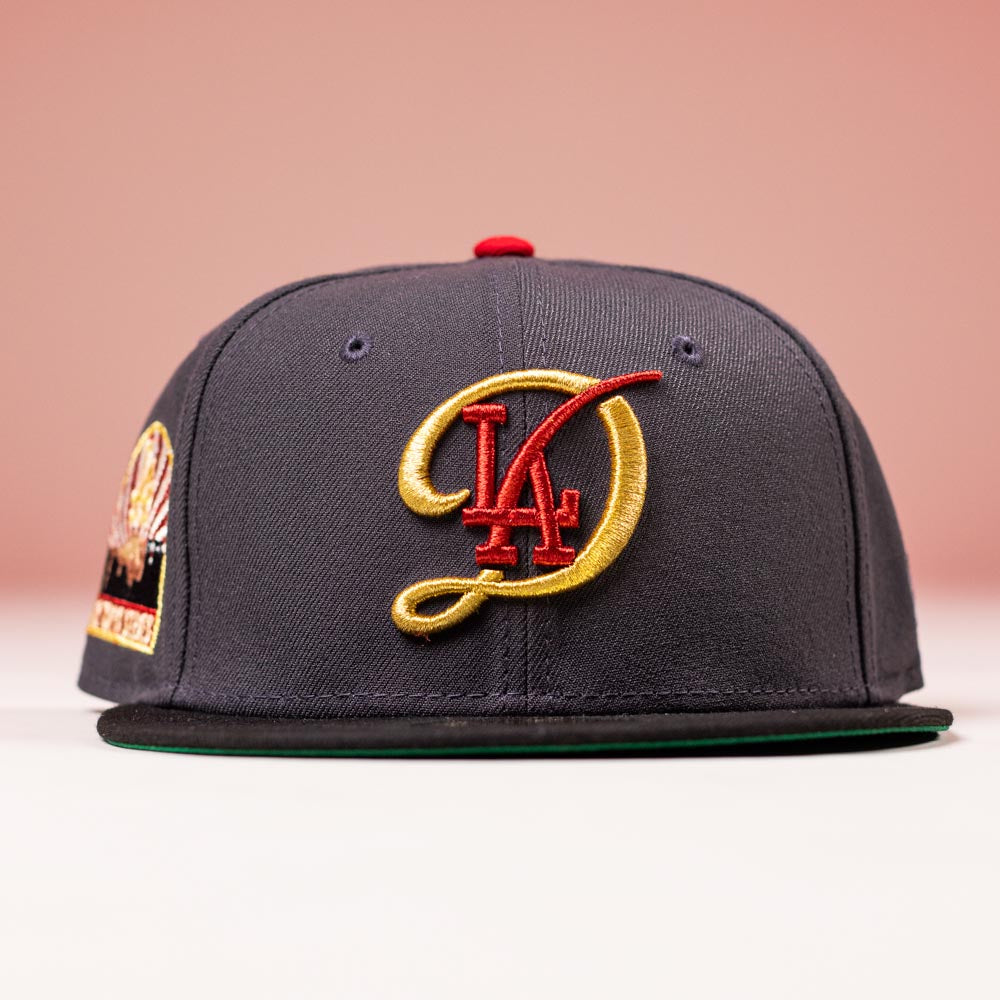 The Bones Pack Los Angeles Dodgers New Era Hat 5950 Fitted features a dark gray design with a gold and red LA logo, red top button, stadium side patch, and is photographed against a pink background—ideal for Dodgers fans.