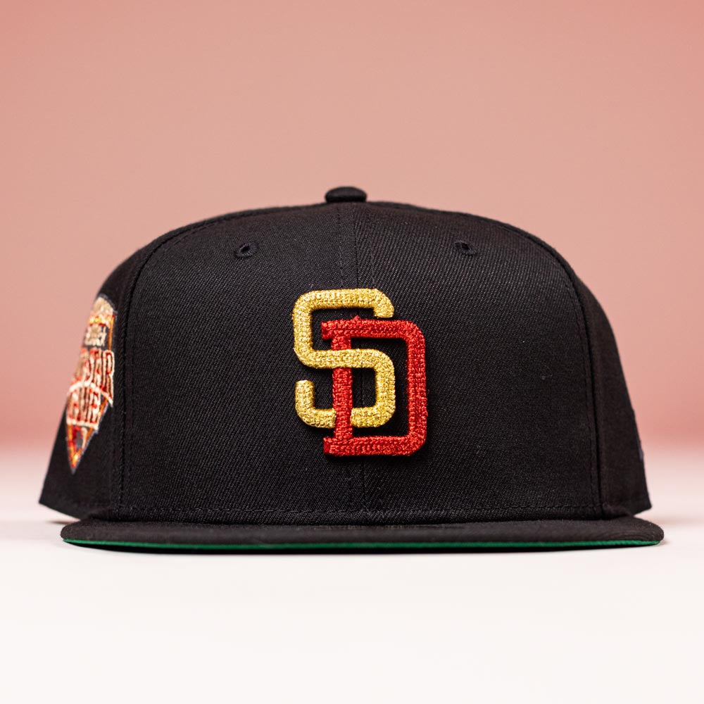 The Bones Pack San Diego Padres New Era Hat 5950 Fitted features a black design with a gold and red SD logo on the front, a colorful patch on the left, and is displayed against a pink background.
