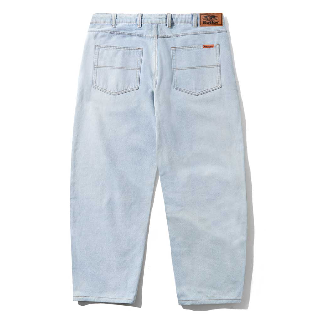 The Baggy Light Blue Denim Jeans by Butter Goods are shown from the back, featuring two rear pockets, a brown leather Butter patch on the waistband, and a red tag on the right pocket. Made from 100% cotton denim, laid flat on a white background.