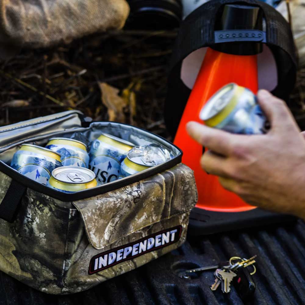 A hand reaches for a can from the Bar Logo 5 Liter Independent x Realtree Cooler Unisex, featuring a camouflage design and INDEPENDENT branding, packed with cans and placed beside an orange traffic cone and some keys—highlighting essential Independent Trucks accessories for any skate session.