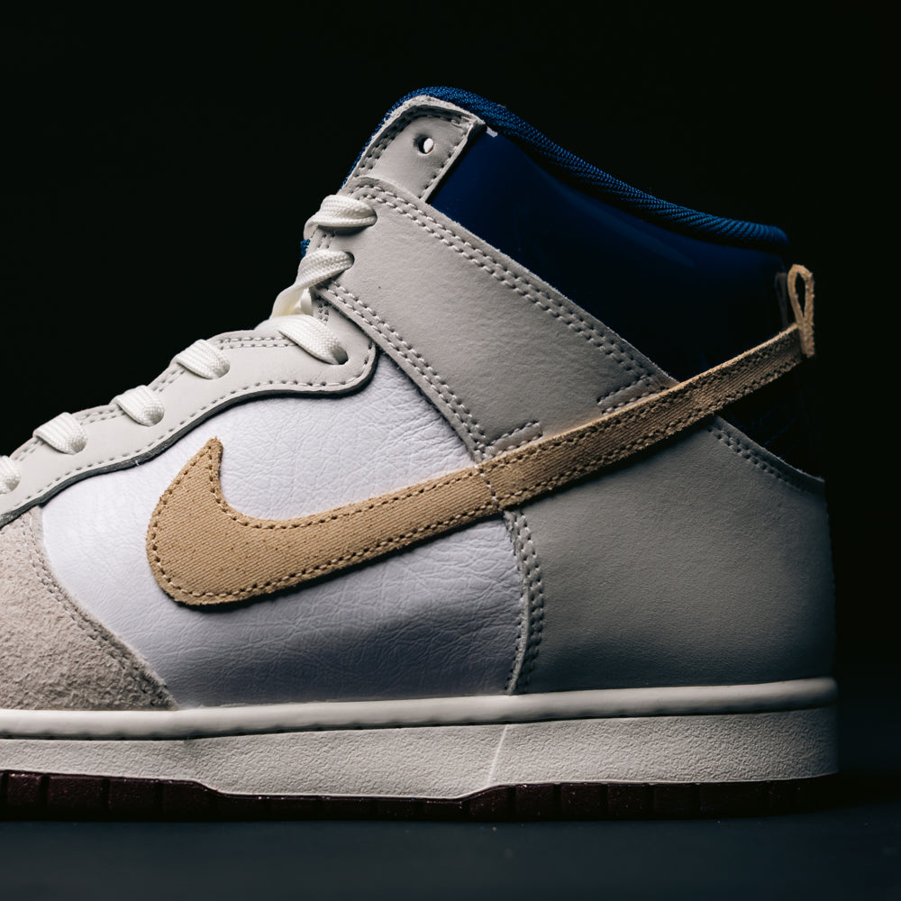 Close-up of the Ivory Sand Blue Void Dunk High Pro x Beyond IU4491-100 Nike SB Skateboarding Shoe, featuring a white and gray upper, beige Nike Swoosh, dark blue heel detail, white laces, and a white midsole against a dark background. This skateboarding sneaker blends classic basketball-inspired style with modern design details.