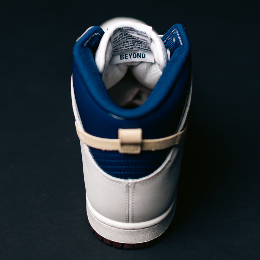 A single Ivory Sand Blue Void Dunk High Pro x Beyond IU4491100 Nike SB Skateboarding Shoe is viewed from the back, featuring a beige strap and the word "BEYOND" visible inside near the heel. The shoe rests on a dark surface with a dark background.