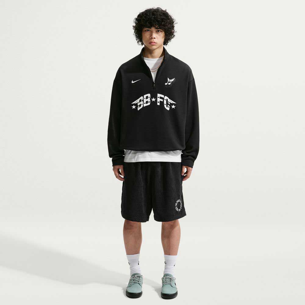 A young person with curly hair stands against a white background wearing the imported black Nike SB Skateboarding IM5463010 Skate 1/2 Zip Fleece Crew, styled with black shorts, white socks, gray sneakers, and featuring white front graphics and easy machine wash care.