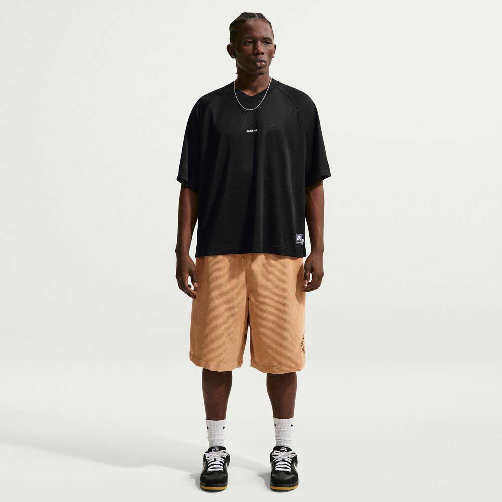 A person stands against a plain white background, wearing the Black Jersey Tee IF1302010 T-Shirt Nike SB Skateboarding Tee, tan knee-length shorts, white socks, black sneakers, with braided short hair and a simple necklace.