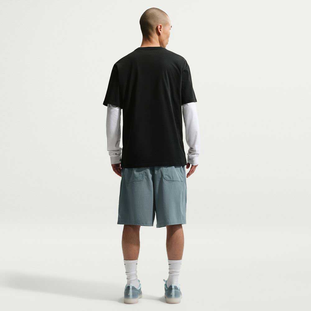 A person stands facing away from the camera, wearing the Black SF Muni Bus Pass Tee IU1265010 T-Shirt Nike SB Skateboarding Tee over a long-sleeve white shirt, light blue shorts, white socks, and light blue sneakers against a plain white background.