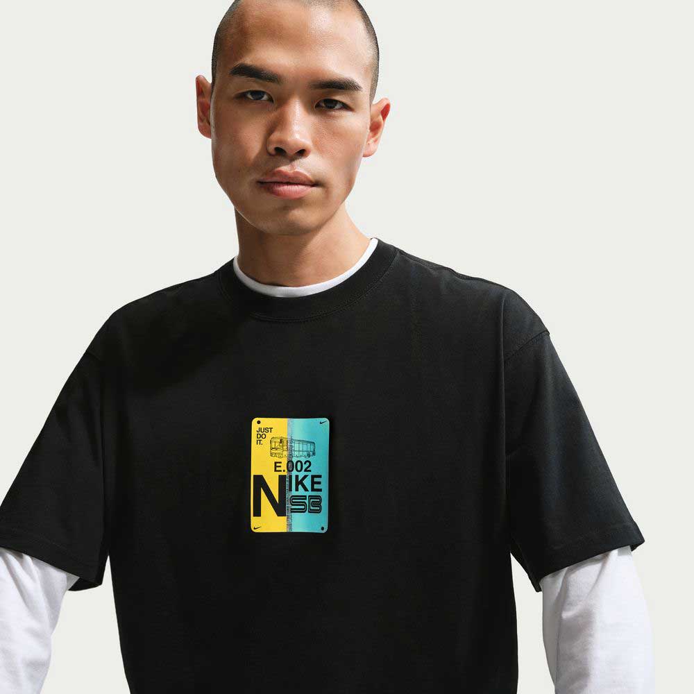Someone wears the Black SF Muni Bus Pass Tee IU1265010 Nike SB Skateboarding T-shirt, a roomy midweight cotton tee with a colorful rectangular graphic and E.002 NIKE N59 text over a white long-sleeve shirt against a plain light background.