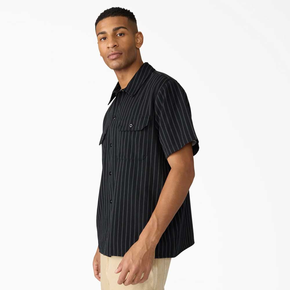 A person wears the Black Worn In Lightweight Work Shirt A89G7BLK Dickies Skateboarding—short-sleeve, relaxed fit, black with white pinstripes—paired with beige pants, standing against a plain white background and looking slightly to the side.