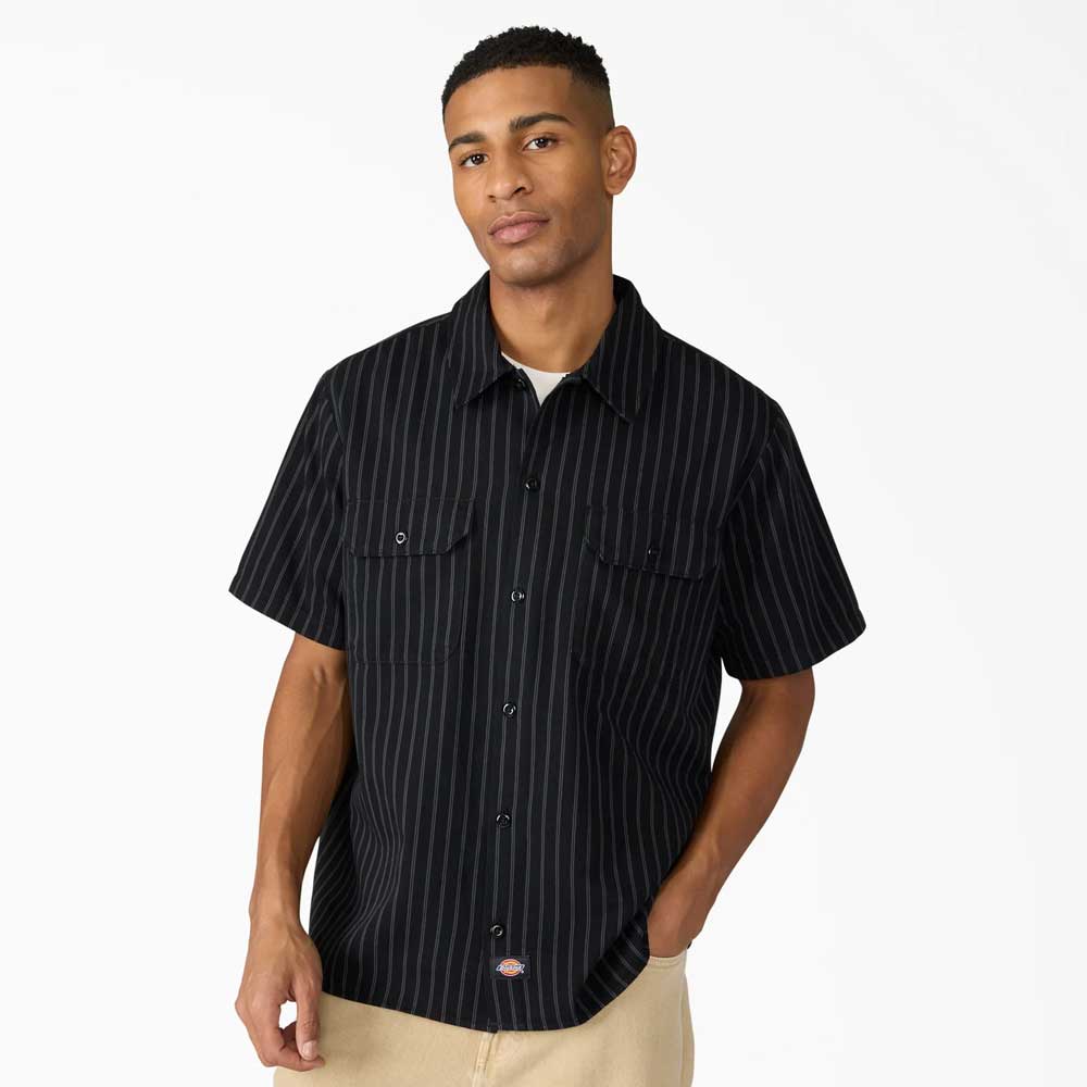 A man wears the Black Worn In Lightweight Work Shirt A89G7BLK by Dickies Skateboarding—100% cotton, black with thin white pinstripes—and tan pants, standing against a white background with one hand in his pocket.