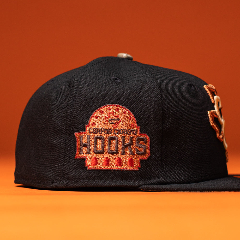 The Hooked in Camo Corpus Christi Hooks New Era Hat 5950 Fitted is a black fitted cap with a red and orange embroidered Corpus Christi Hooks logo on the side, displayed against an orange background.