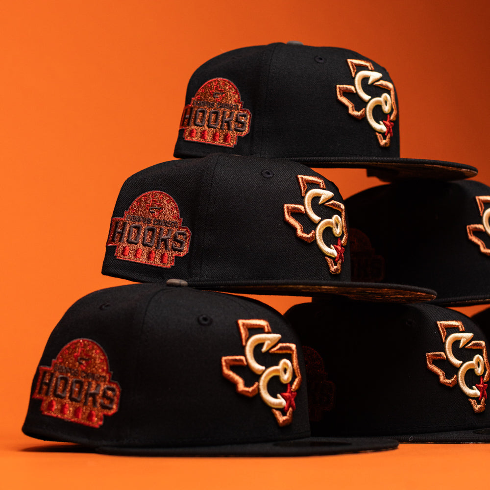Six Hooked in Camo Corpus Christi Hooks New Era Hat 5950 Fitted caps are stacked in two uneven columns against an orange background. Each black cap features bold orange and red embroidered logos—including a Texas state outline and the word HOOKS—showcasing the distinctive style of the Corpus Christi Hooks.