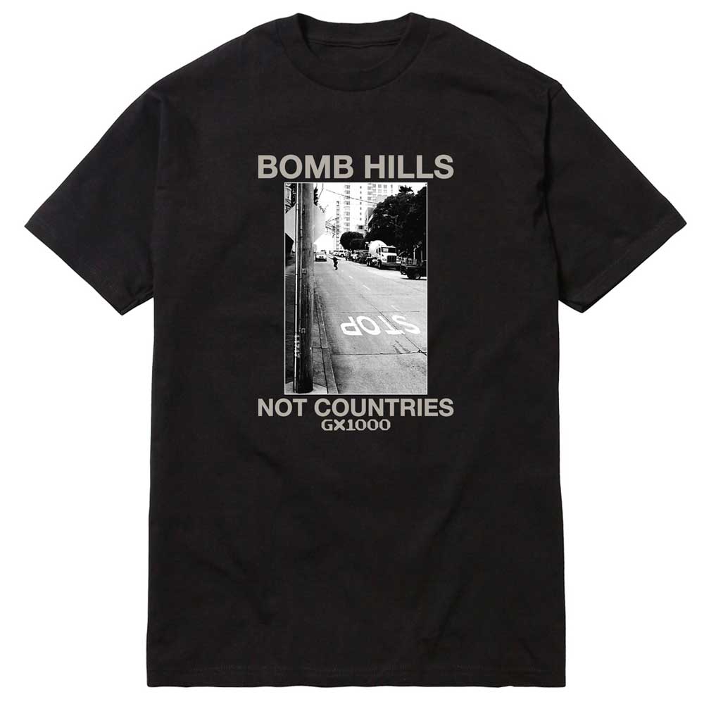 The Bomb Hills Not Countries Black GX1000 Tee Gray Font showcases a striking black-and-white photo of a street hill with a prominent STOP road marking. The gray font above the image reads “BOMB HILLS,” while the text below states “NOT COUNTRIES,” making this tee a standout addition to any collection.