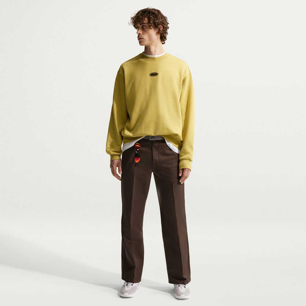 A person with curly hair stands against a plain white background, wearing the Buff Gold Crew Fleece Nike SB II4440720, brown pants, white sneakers, and a keychain clipped to their pants.
