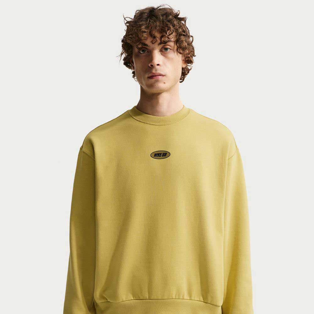 A young man with curly hair wears the Buff Gold Crew Fleece Nike SB II4440720, featuring a small black and white oval logo on the chest, standing against a plain white background.