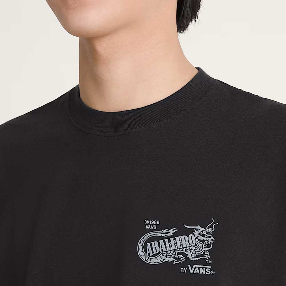 Someone wearing the Caballero VN000XHBBLK1 Black Short Sleeve Tee from Vans Skateboarding, featuring a small dragon graphic and "CABALLERO BY VANS" text with copyright 1989 on the chest; only their lower face and neck show.