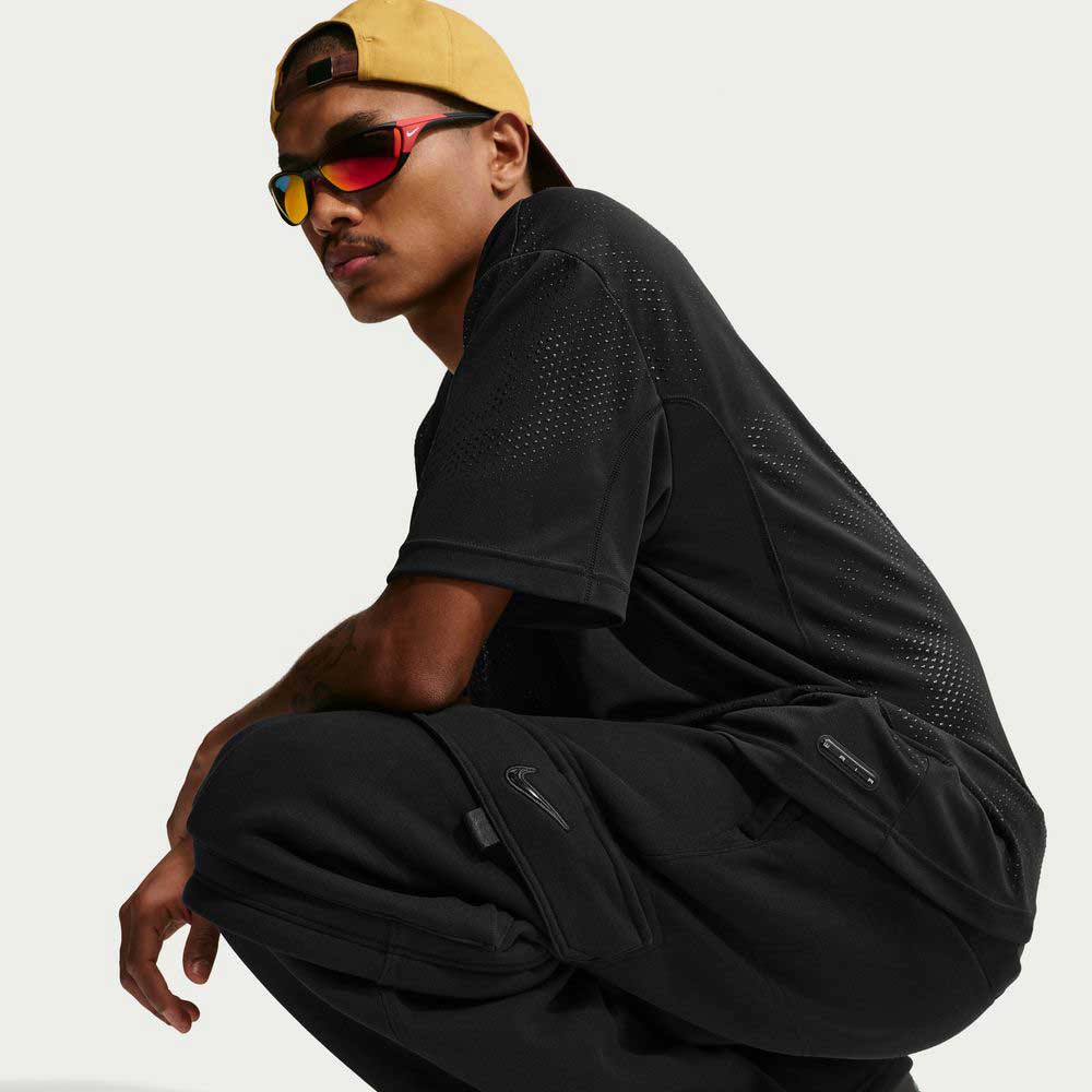 A young man squats in a relaxed pose, wearing Nike SB Ishod Cargo Pant Sweatpant in black, paired with a yellow backwards cap and reflective sunglasses. A black Nike logo is visible. The background is plain and light-colored.