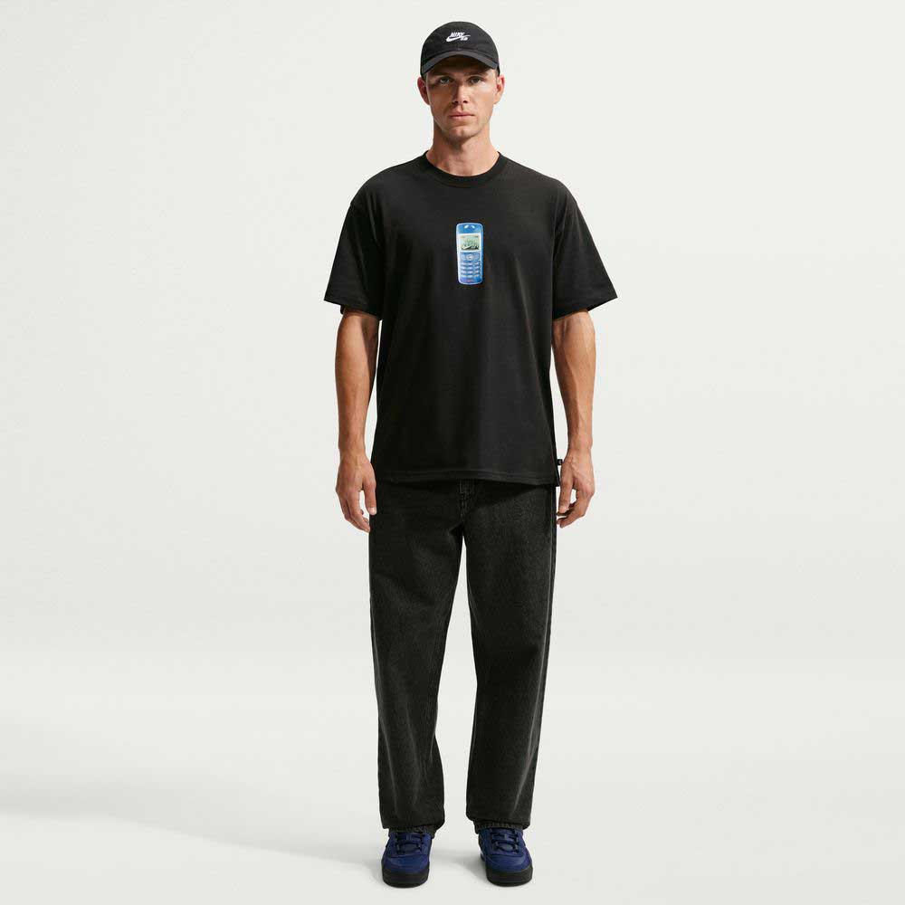 A man wearing a black cap, the Nike SB Skateboarding Cell Phone Tee Black IM4424010 T-shirt with a small chest graphic, loose black jeans, and dark sneakers stands facing forward against a plain white background.