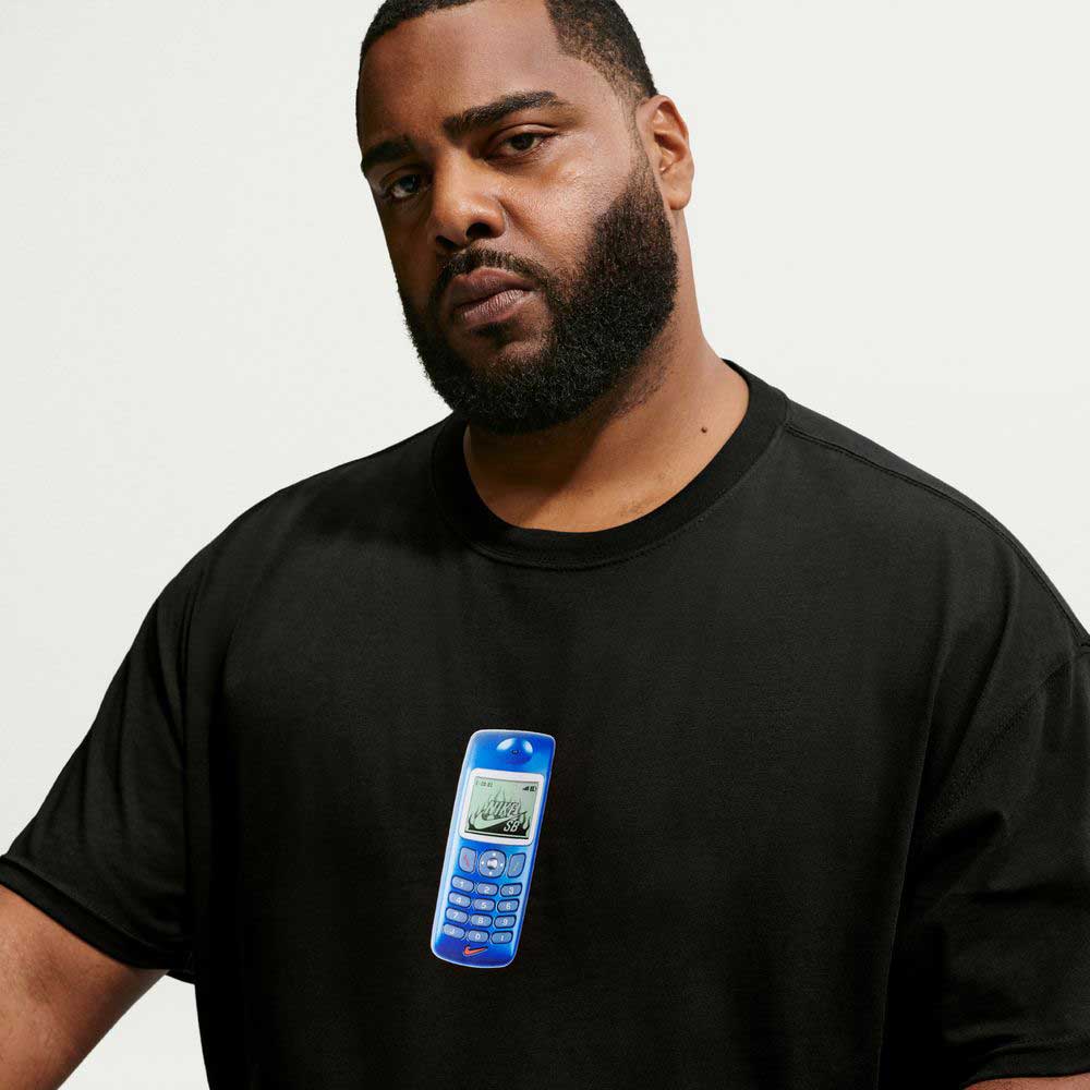 A man with a beard wears the Nike SB Skateboarding Cell Phone Tee Black IM4424010 T-Shirt, which features a blue retro mobile phone graphic on the front. Reminiscent of the comfort found in a Nike SB Jacquard Skate Jersey, this tee combines casual style with the feel of your favorite skate jersey top. The background is plain white.