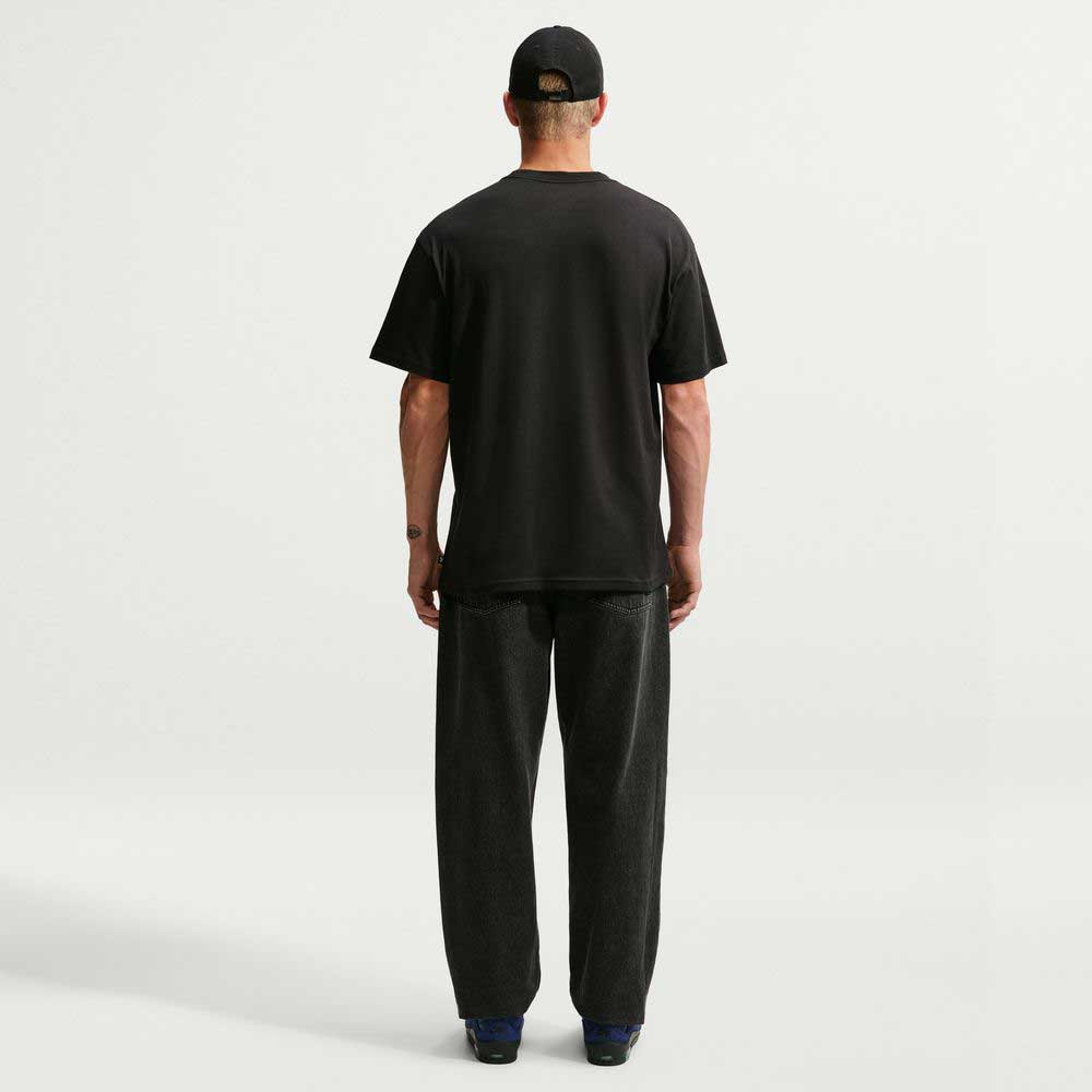 A person stands facing away, wearing a black Nike SB Cell Phone Tee (IM4424010) paired with dark loose pants, a black cap, and dark shoes, on a plain white background. The sweat-wicking Nike SB Skateboarding t-shirt combines comfort and style for an effortlessly cool look.