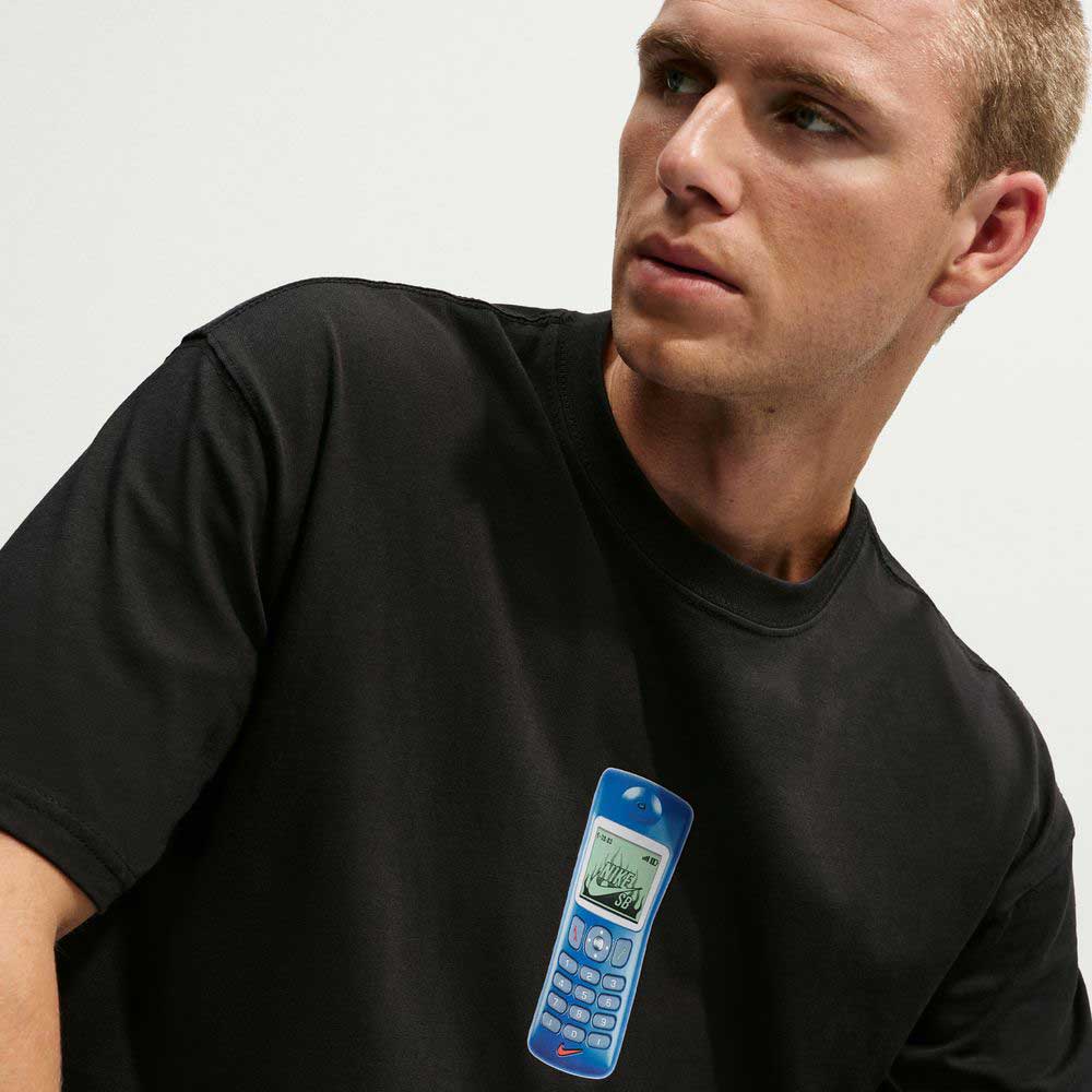 A person wearing a black Nike SB Cell Phone Tee (IM4424010) featuring an image of a blue toy phone looks slightly to the side. This sweat-wicking skateboarding T-shirt blends comfort with playful style.