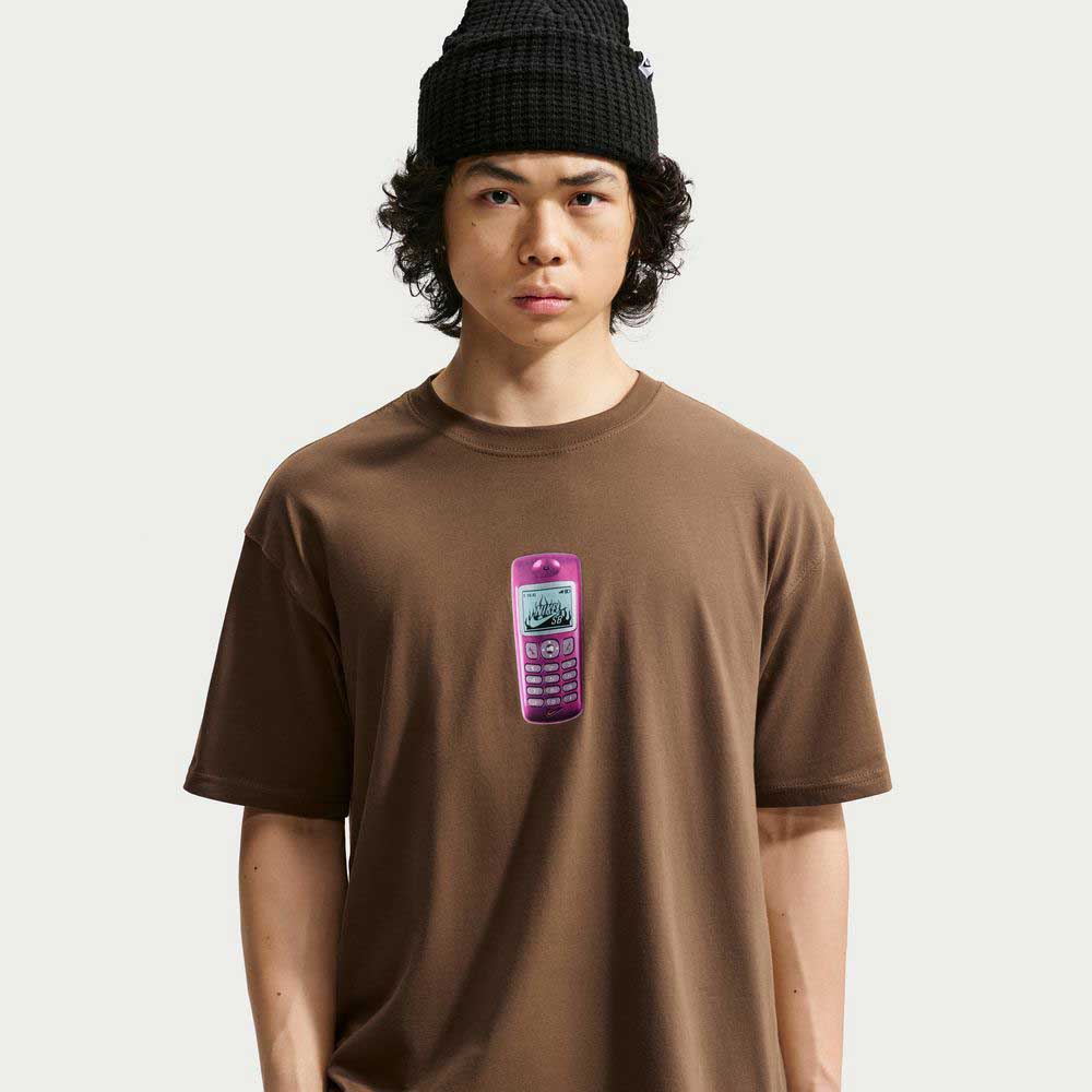 A person wearing a black knit beanie and the Nike SB Skateboarding Cell Phone Tee Brown IM4424235, featuring a graphic of a pink vintage mobile phone, stands against a plain light background, their outfit completed by the sweat-wicking comfort of the Nike SB Jacquard Skate Jersey layered as an effortlessly cool skateboarding top.