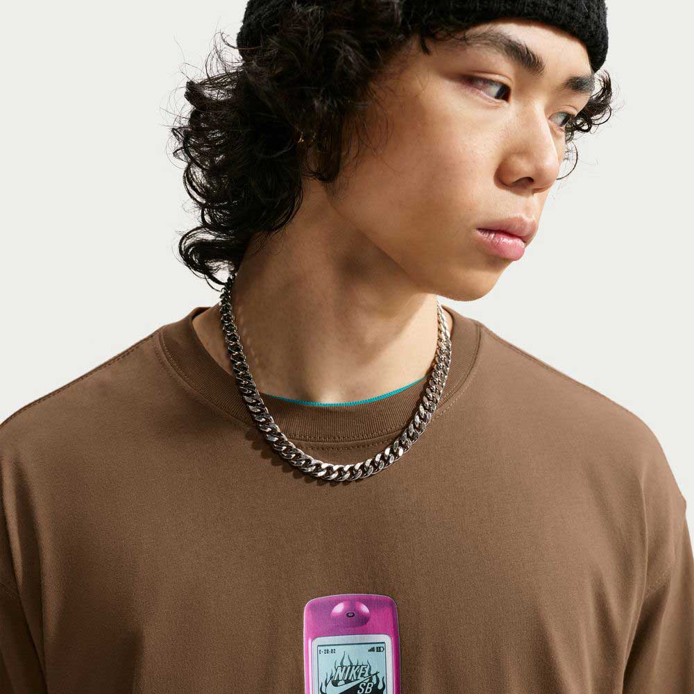 A young person with curly hair wears a black beanie, two silver chains, and the Nike SB Cell Phone Tee in Brown (IM4424235), an essential skateboarding t-shirt. Looking to the side, they have a pink flip phone with a sticker partially visible hanging from their neck.