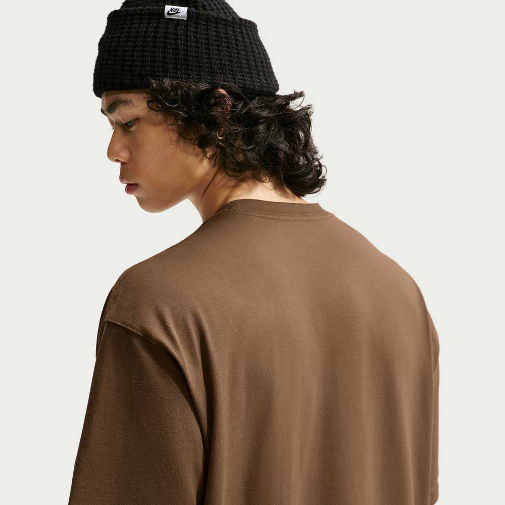 A person with curly hair, seen in side profile and turned away from the camera against a plain background, is wearing a black knit beanie and the Nike SB Skateboarding Cell Phone Tee Brown IM4424235 t-shirt.
