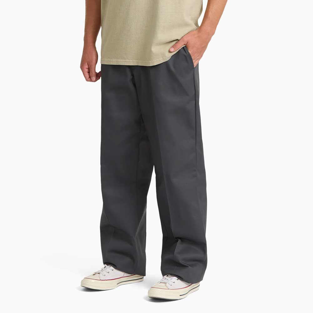 A person wearing Charcoal Loose Fit Work Pant A88KB0CH Dickies Skateboarding, a beige shirt, and white sneakers stands with hands in pockets against a plain white background.