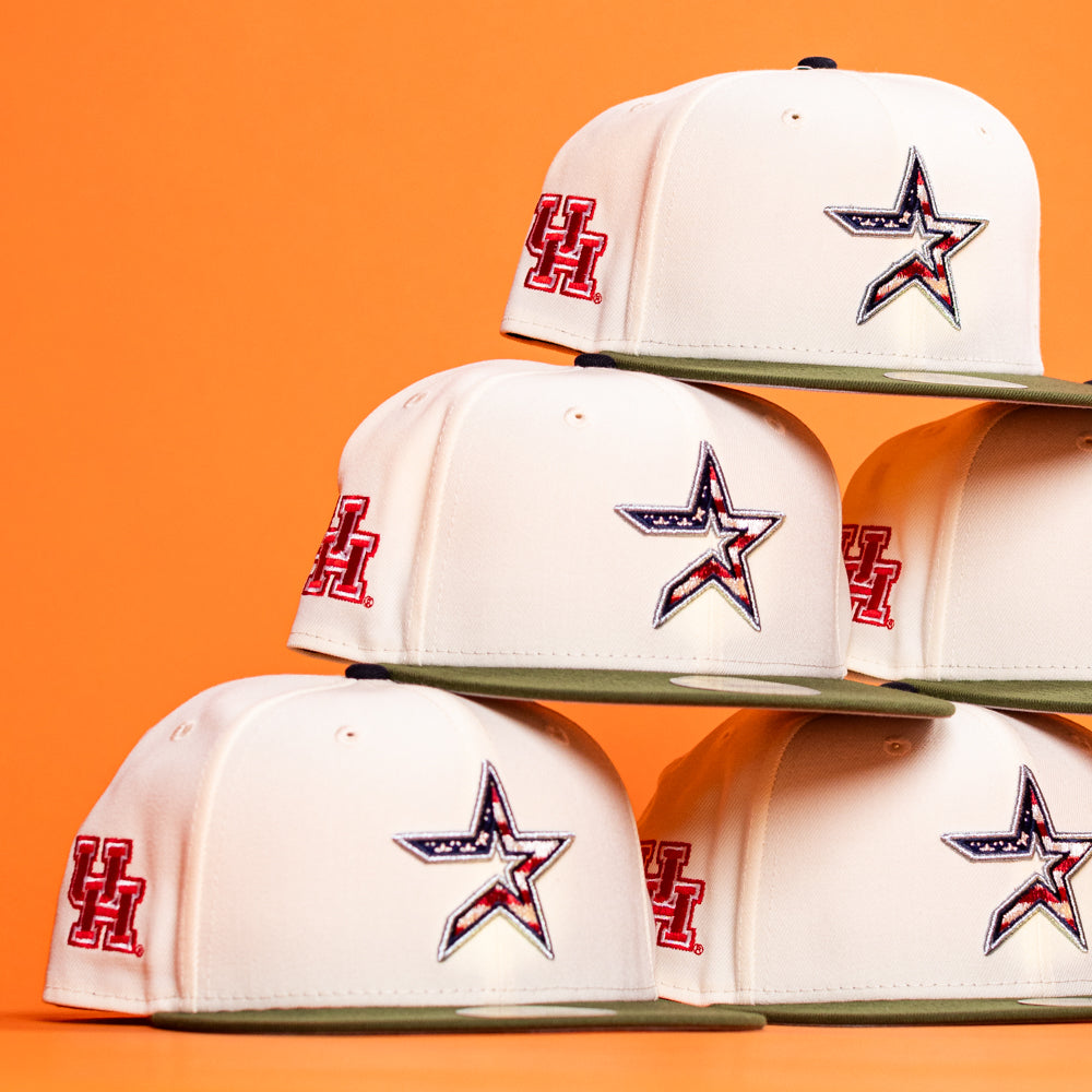 A stack of Stars Over Houston UH New Era Hat 5950 Fitted hats in white with olive brims, a red UH logo on the side, and a front star outlined in black with an American flag pattern—all on an orange background.