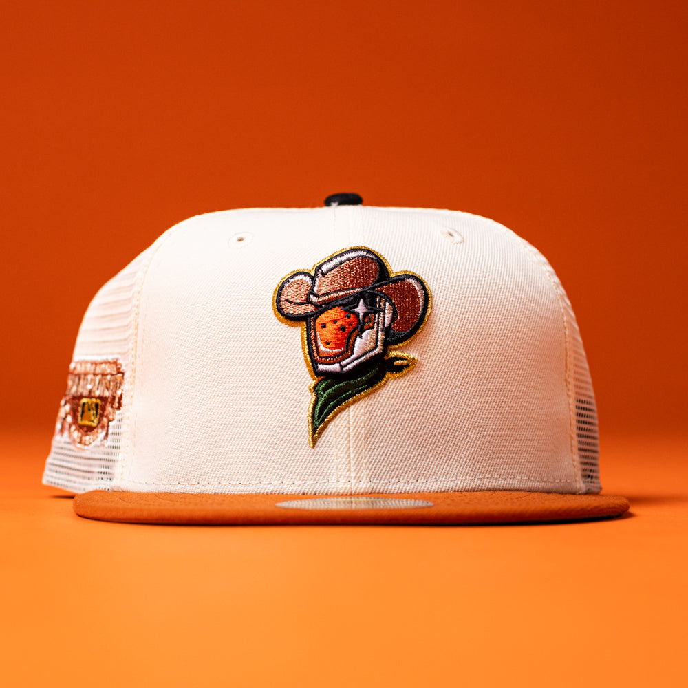 The Wrangler Chrome Rust Sugarland Space Cowboys New Era Hat 5950 Fitted, a white and orange trucker hat featuring an embroidered patch of a cowboy-themed orange on the front, from the Space Cowboys Hat collection, is displayed against an orange background.
